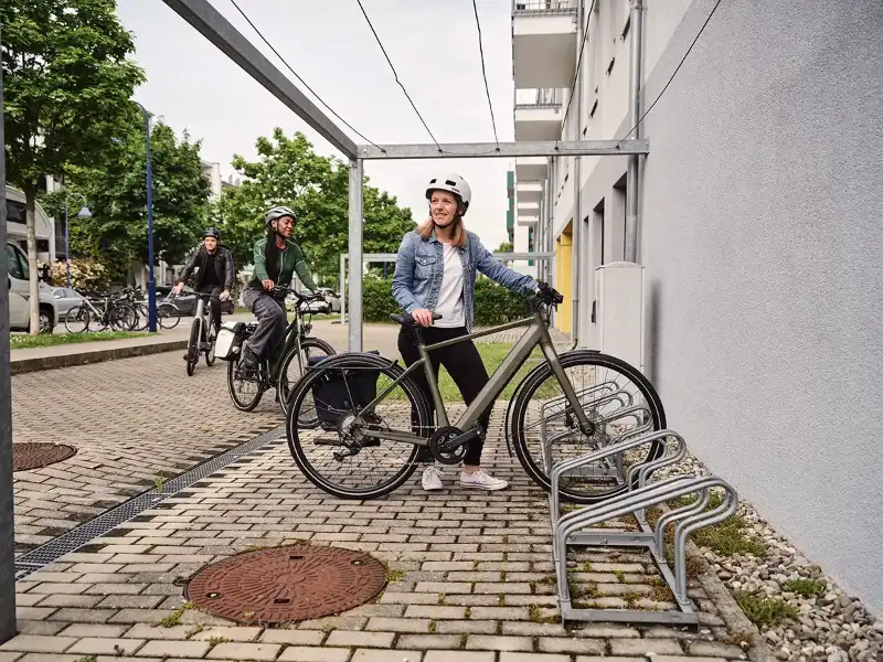 Eine Frau in blauer Jeansjacke parkt ihr grünes E-Bike, eine weitere Frau und ein Mann fahren den Fahrradstellplatz an