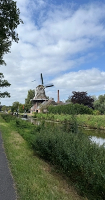 Eine traditionelle Windmühle steht neben einem kleinen Kanal, umgeben von grünen Wiesen und Bäumen unter teils bewölktem Himmel.